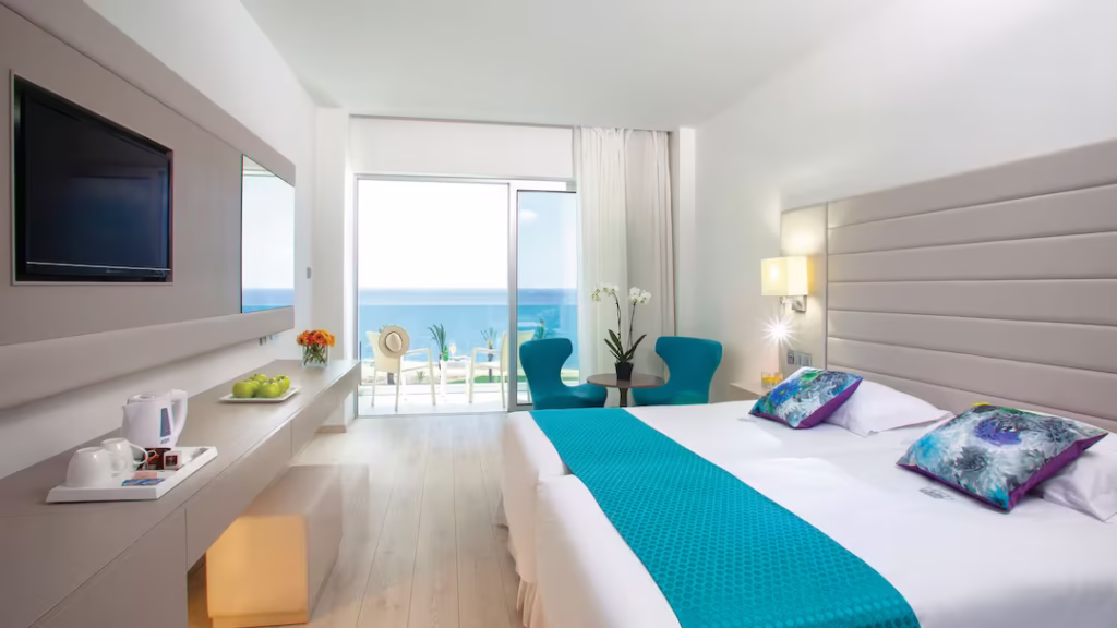 Bright hotel room at King Evelthon Beach Hotel & Resort with large window overlooking the sparkling Mediterranean Sea and sunlit balcony.