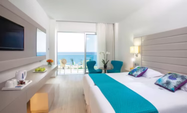 Bright hotel room at King Evelthon Beach Hotel & Resort with large window overlooking the sparkling Mediterranean Sea and sunlit balcony.
