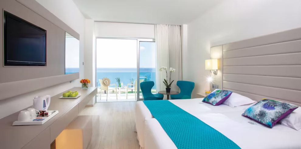 Quarto de hotel luminoso no King Evelthon Beach Hotel & Resort, com uma grande janela com vista para o cintilante Mar Mediterrâneo e uma varanda iluminada pelo sol.